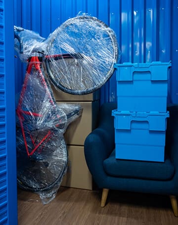 Shrink-wrapped bicycle stored beside boxes and an armchair in a blue storage unit